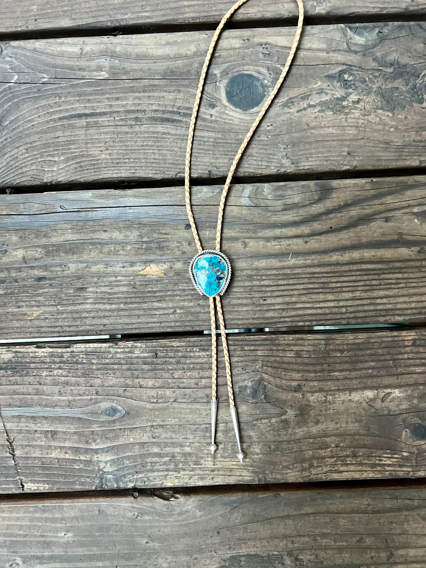 Bolo tie with shattuckite chrysocolla set in sterling silver and tan leather cord. Handmade bolo tie using metal smith techniques. Displayed on wooden background.