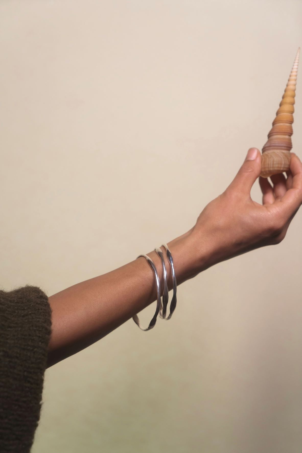 Handcrafted sculpted bangles, this creation by Soulfull Studio is a product of intricate artistry. Utilizing lost wax casting techniques with recycled silver, it derives its inspiration from the allure of natural elements. Jewelry for the wild ones. Model with beige background holding shell.