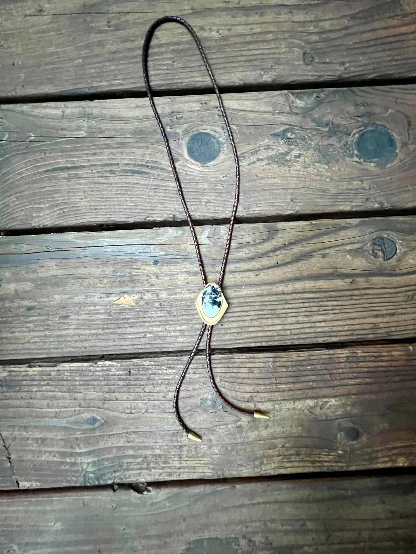 Bolo tie with white buffalo set in brass and brown leather cord. Handmade bolo tie using metal smith techniques. Displayed on wooden background. 