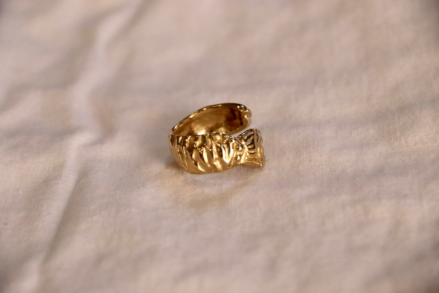 A textured yellow brass fish rings, sculpted  to show scales and tail. Resting on a soft, off-white fabric background.