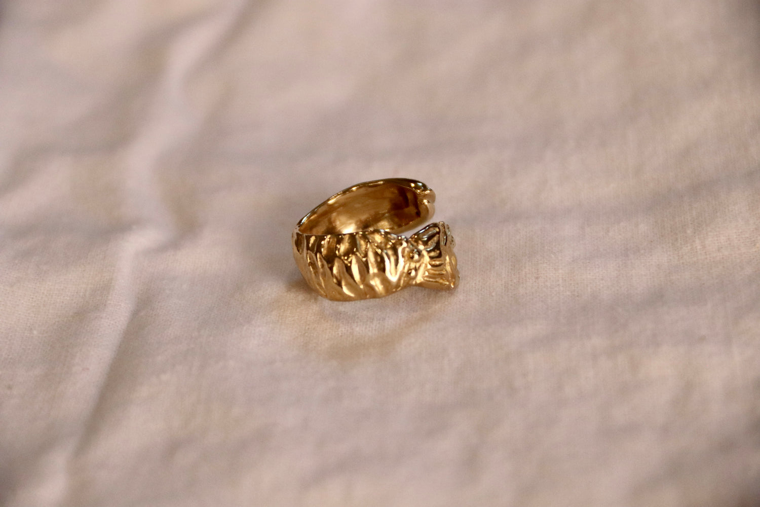 A textured yellow brass fish rings, sculpted  to show scales and tail. Resting on a soft, off-white fabric background.
