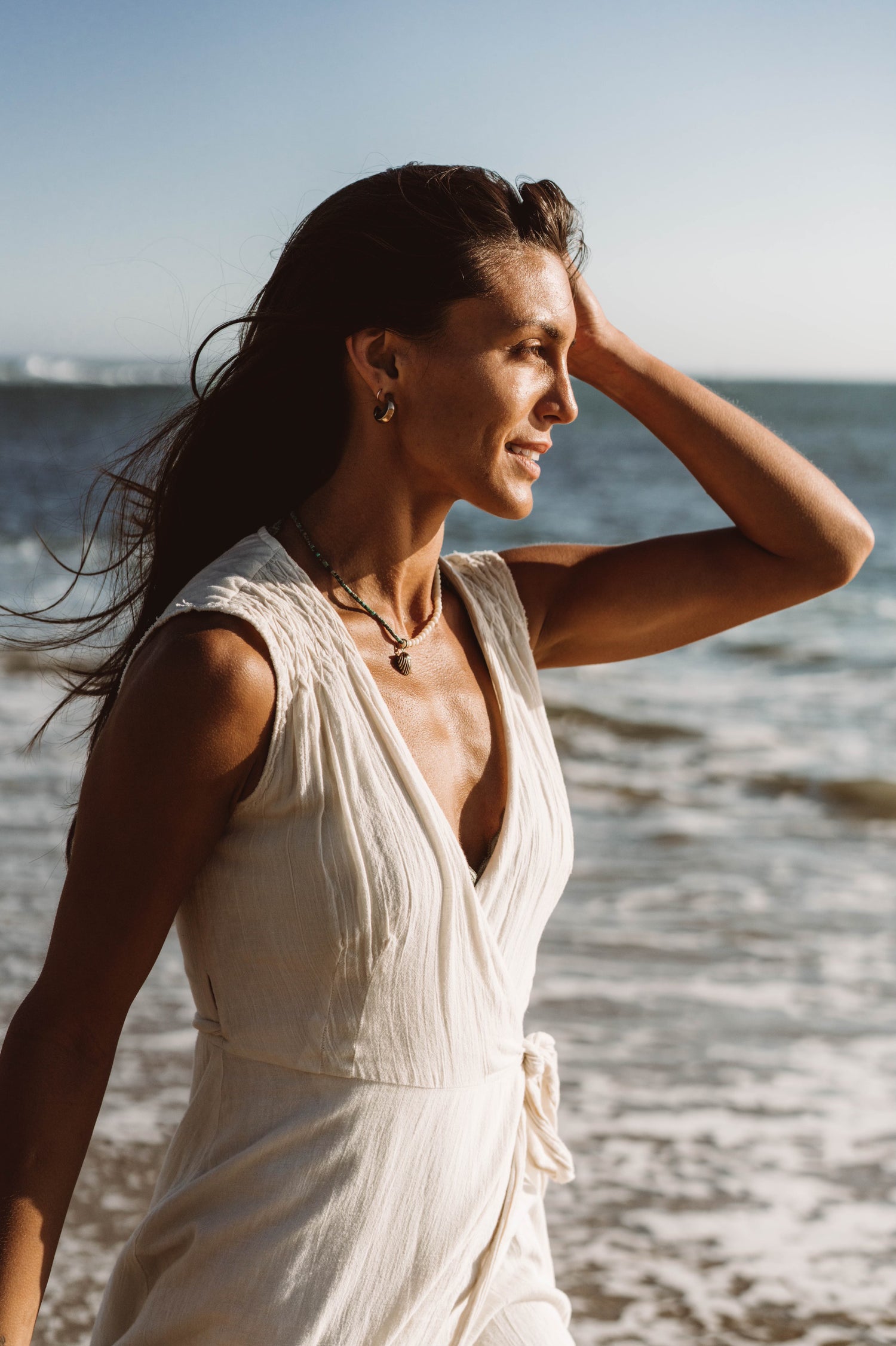 Land & Sea necklace. Made with bronze pendant, turquoise beads, and coral beads. Model in white dress at beach. 